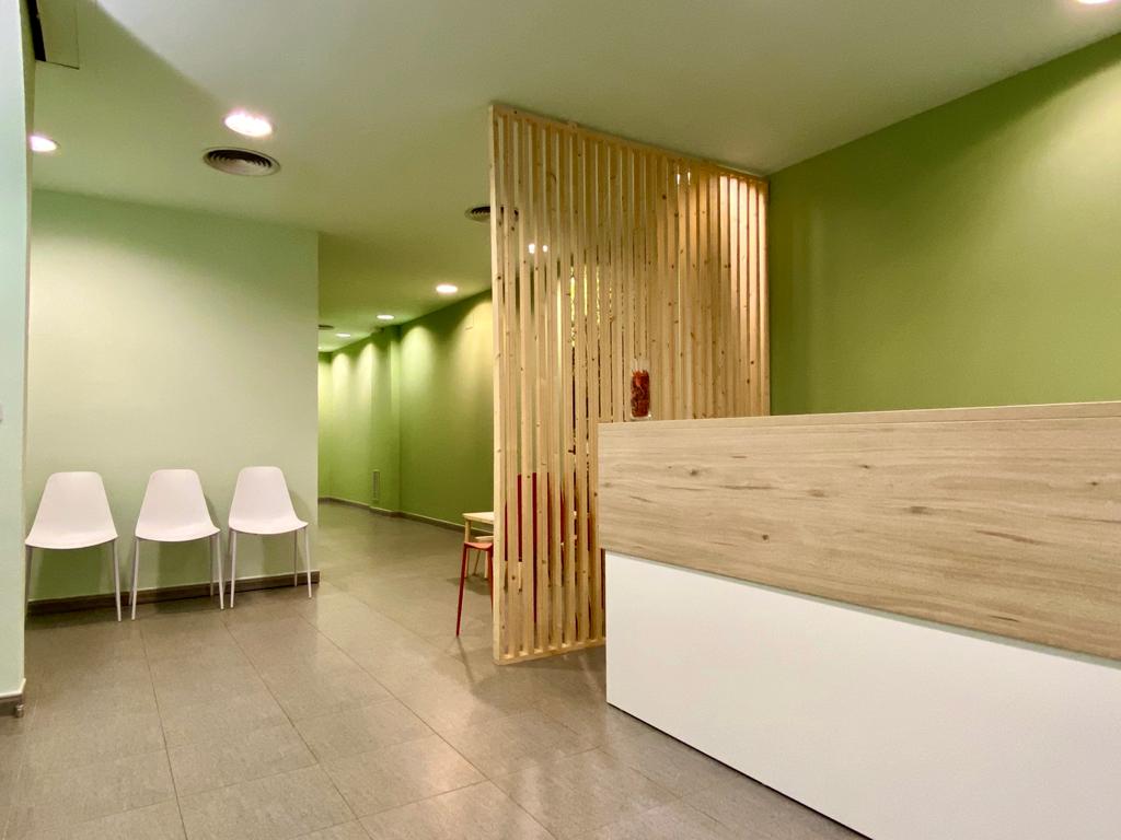Pasillo y recepción de una clínica: paredes verde pistacho iluminadas por focos, celosía de listones de pino, mostrador con tablero de roble claro sobre base blanca, tres sillas blancas de una pieza y suelo de baldosa gris.